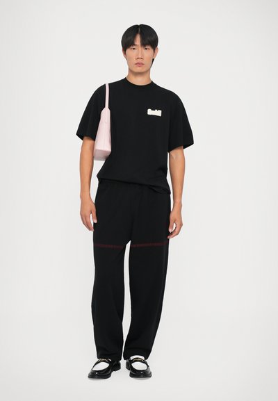 Zwarte T-shirt met een wit logo, gecombineerd met zwarte broek met rode stiksel-details. Het model draagt een lichtroze handtas en heeft zwarte schoenen aan.