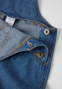 Blauwe denimstof met een lichte wassing, met zichtbaar stikken, metalen drukkers en een verzorgingslabel. Het ontwerp bevat een omgeslagen kraag en een zak.