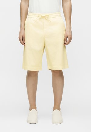 Mężczyzna w bladożółtych szortach z troczkiem, białych butach wsuwanych i białej bluzce, stojący na jednolitym tle.