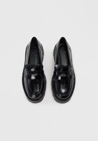 Paar mustade läikivate nahast DKNY mokassiinide brogue-detailidega ja paksude kummitaldadega, vaadatuna ülalt valge taustal.