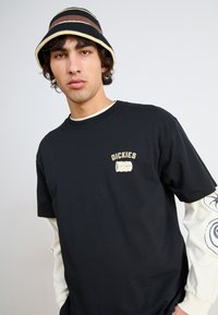 T-shirt en coton noir avec un logo "Dickies" en beige. Superposé avec une chemise blanche à manches longues ornée de motifs bleus. Chapeau seau noir et marron à rayures.