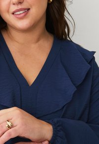 Blusa blu navy con scollatura a V, caratterizzata da un colletto arricciato e polsini elasticizzati. Il modello indossa un anello d'oro sulla mano sinistra. Tessuto strutturato.