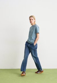Personne aux cheveux blonds portant un t-shirt bleu, un jean bleu ample et des baskets marron marchant sur un sol vert contre un mur blanc.
