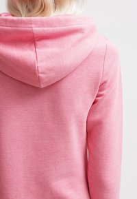 Sweat à capuche rose en tissu doux et texturé, avec un cordon de serrage à la capuche et des détails cousus.