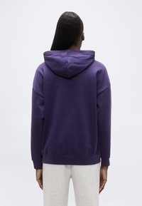 Μωβ φούτερ με κουκούλα από μείγμα βαμβακιού, με άνετη εφαρμογή και ριμπ μανσέτες, σε συνδυασμό με γκρί φόρμα. Εμφάνιση από πίσω.