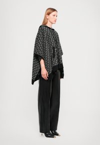 Knitted poncho in black and gray with geometric pattern, features a hood, zip closure, and two pockets. Worn over black wide-leg trousers.