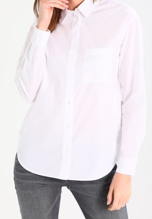 Vrouw draagt een wit hemd met lange mouwen en knopen, met een borstzak en donkergrijze spijkerbroek, hand bij haar kin en heup.