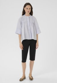 Blusa azzurro chiaro, con forma quadrata e pieghe, scollatura tonda e maniche corte; abbinata a pantaloni neri fino al ginocchio e ballerine marroni.