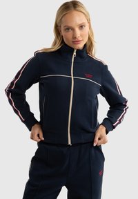 Veste de survêtement navy avec un col haut, des bandes latérales blanches et rouges, une fermeture éclair, et des détails de logo rouges. Tissu texturé.