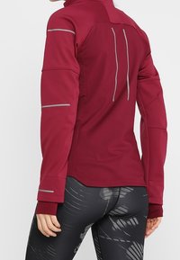Bordowa sportowa kurtka z długimi rękawami i odblaskowymi akcentami; posiada wysoki kołnierz i dopasowany krój. Teksturowany materiał, elegancki design.