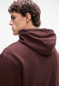 Brun hettegenser med stor hette, laget av mykt stoff. Har et enkelt design med sømdetaljer langs sømmene.