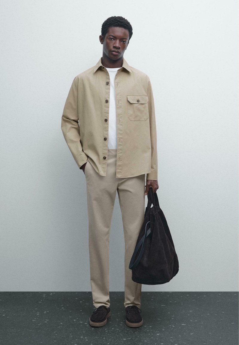 Beige cotton shirt with buttons, white t-shirt, beige trousers, and dark suede bag. Model stands on textured dark floor with light backdrop.