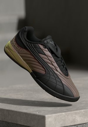 Must musta ja pruuni Puma sneaker kollase tallaga, hõljuv betoonserva kohal tumeda udustes taustal.