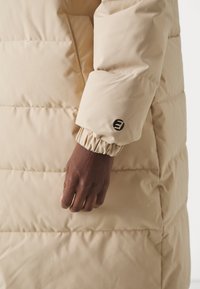 Manteau doudoune beige avec des sections matelassées. Présente un poignet froncé et un logo noir sur la manche. Texture de tissu lisse et mate.