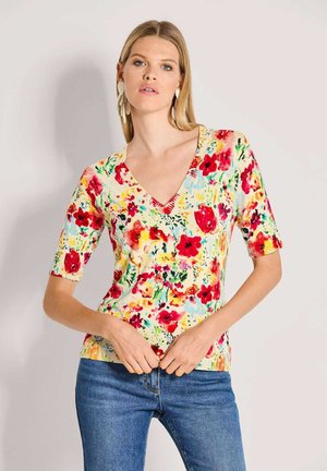 Vrouw met grote witte oorbellen, een kleurrijke V-hals trui met bloemenmotief en halflange mouwen, en blauwe spijkerbroek, staand tegen een lichte achtergrond.