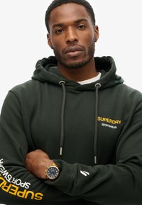 Felpa verde scuro in cotone, con un logo frontale giallo, cordini regolabili e un orologio con quadrante blu.