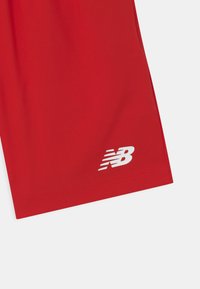 Punaiset urheilushortsit, jotka on valmistettu kevyestä kankaasta. Shortseissa on valkoinen "NB"-logo vasemmassa alakulmassa ja sileä pintakuvio.