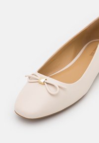 Chaussure plate pour femme couleur crème avec bout arrondi, dotée d'un petit nœud décoratif avec un accent doré sur le dessus.