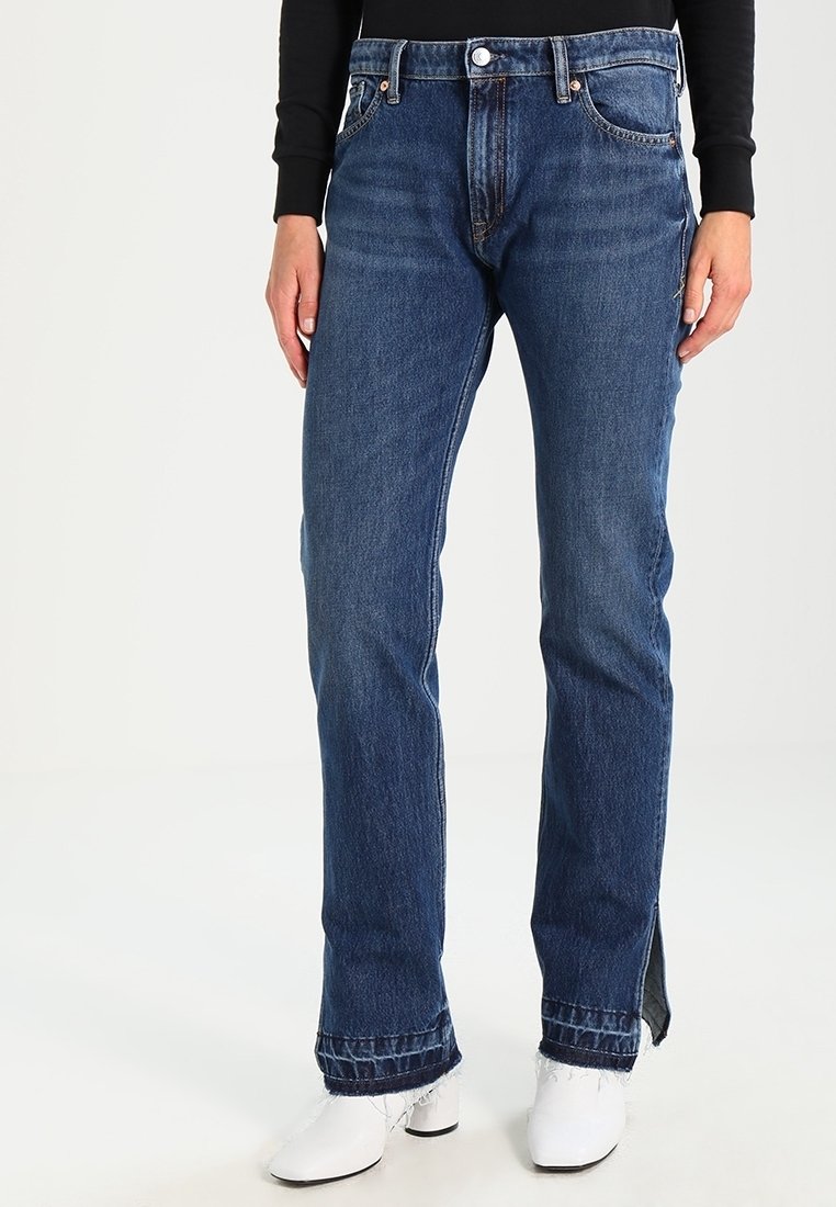 Jeans de denim azul con un corte recto, cinco bolsillos, cierre de botón y detalles de costura sutiles. El dobladillo presenta acentos divididos en ambos lados.