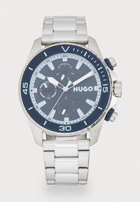 Chronograph watch with a silver stainless steel case, blue dial, and white bracelet. Features date, weekday, and sub-dials. Blue accents on bezel.