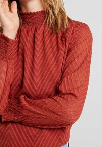 Röd genomskinlig blus med hög smockad halsringning, med ett zigzag-mönster och samlade ärmslut. Lätt och flödande tyg.