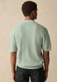 Short-sleeve polo in mint green textured knit with a ribbed collar and hem, paired with dark denim jeans. Back view showcases fit and design.
