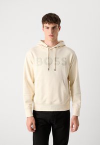 Sudadera color crema con un gran logo de BOSS en relieve en la parte delantera, capucha ajustable con cordón y bolsillo tipo canguro. Combinada con pantalones negros.