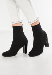 Czarne botki sięgające kostki, wykonane z teksturowanej dzianiny, z wysokim słupkowym obcasem i wycięciami po bokach. Gładki czubek i nowoczesna sylwetka.