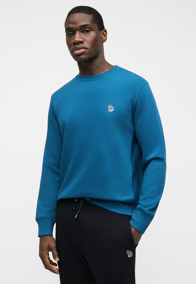 Άνδρας που φοράει μπλε φούτερ μακρυμάνικο και μαύρο παντελόνι, και τα δύο με μικρά πολύχρωμα λογότυπα ζέβρας, στέκεται μπροστά σε απλό φόντο.