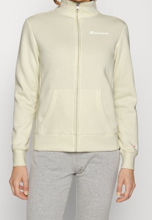 Béžová fleecová bunda Champion na zip s vysokým límcem, předními kapsami a logem na hrudi a manžetě rukávu, nošená s šedými kalhotami.