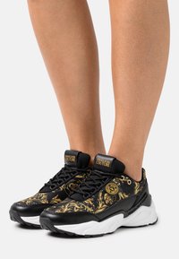 Baskets noires avec un motif floral doré, dotées d'une semelle rembourrée, d'un design à lacets et d'accents de logo sur la languette et sur le côté.