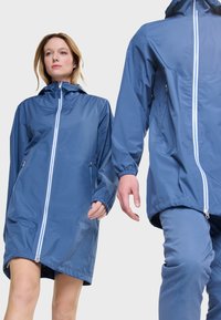 Mujer y hombre con chaquetas impermeables azules a juego con cremalleras delanteras, la chaqueta de la mujer llega hasta la mitad del muslo, de pie frente a un fondo blanco.