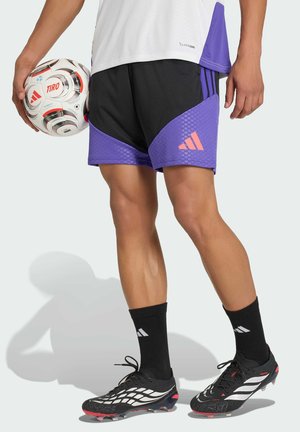 Osoba ubrana w czarne i fioletowe krótkie spodenki Adidas, czarne skarpetki i korki, trzymająca biało-czerwoną piłkę do piłki nożnej Adidas TIRO przy boku.