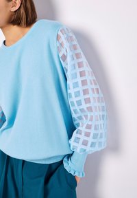 Hellblauer Pullover mit einem einfarbigen Körper und durchsichtigen, karierten Ärmeln; weiche Textur und gerippte Bündchen. Kombiniert mit petrolfarbenen Hosen.
