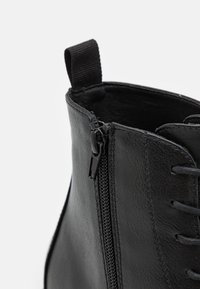 Bottine en cuir noir avec fermeture éclair latérale, lacets et finition texturée. Comprend une languette en tissu pour faciliter l'enfilage.