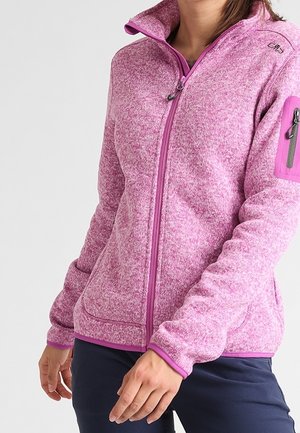 Vrouw draagt een roze fleecevest met rits en een zak op de linker mouw, samen met een donkere broek, staand tegen een lichte achtergrond.