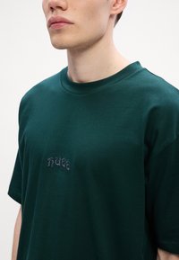 Mørkegrønn bomulls T-skjorte med rund hals, med brodert tekst på brystet. Korte ermer og en glatt tekstur.