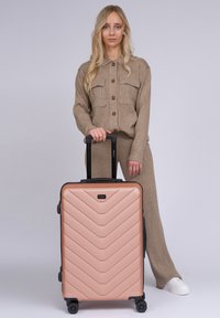 Roségoldener Koffer mit Chevron-Muster, schwarzen Akzenten und einem ausziehbaren Griff. Verfügt über vier Räder für Mobilität.
