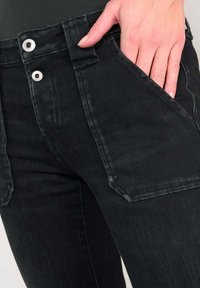 Jeans de denim negro de cintura alta, con dos bolsillos frontales, herrajes de botón plateado y una textura desgastada.