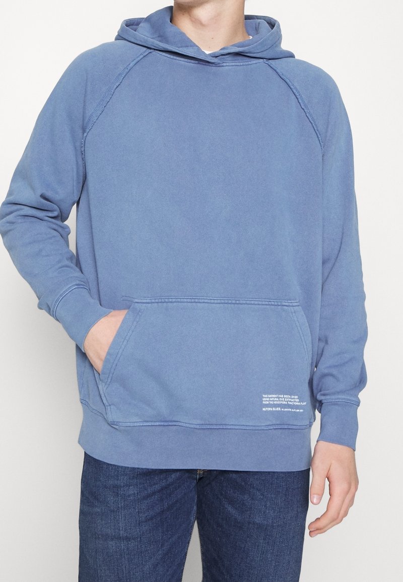 Homme portant un sweat à capuche bleu avec une poche avant et un petit texte blanc près de la poche, associé à un jean bleu foncé, sur fond uni.