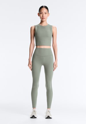 Ženska stoji obrnjen naprej, oblečena v ujemajočo se svetlo zeleno brezrokavnik in kratko majico ter hlačne pajkice z visokim pasom, obuta v bele superge, na belem ozadju.