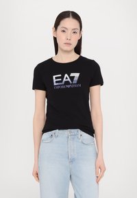 Zwarte katoenen t-shirt met het "EA7 Emporio Armani" logo in metallic zilver en blauw. Korte mouwen, ronde hals, slim fit. Gecombineerd met lichtblauwe spijkerbroek.