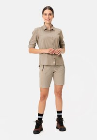 Camicia beige con bottoni e maniche arrotolate, abbinata a pantaloni corti beige e scarpe da trekking nere. Presenta tasche e calze nere a righe.