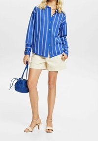 Camisa de botones a rayas azules, pantalones cortos crema, sandalias de tacón beige y un pequeño bolso tejido azul. Tela suave y corte relajado.