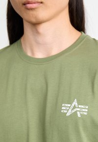 Olivenfarvet T-shirt lavet af bomuld, med en hvid grafisk logo på venstre bryst, rund halsudskæring og korte ærmer.