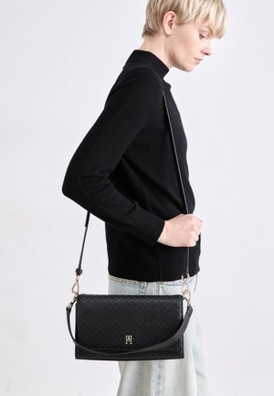 Personne portant un pull noir et un jean clair tient un sac bandoulière noir texturé avec des détails en métal doré et un logo.