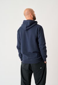 Navyblauer Hoodie aus weichem Stoff, mit Kordelzugkapuze, gerippten Bündchen und Saum. Mit schwarzen Hosen getragen, von hinten betrachtet.