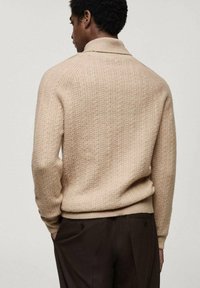 Pull en tricot beige avec un motif texturé, présentant un col haut et des poignets côtelés. Le dos affiche une coupe ajustée et un tissu doux.