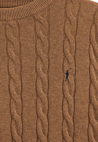 Brauner Strickpullover mit Zopfmuster, ausgestattet mit einem gerippten Ausschnitt und einem kleinen marineblauen Logo, das vorne aufgestickt ist.