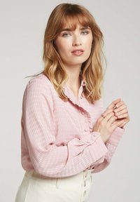 Blouse boutonnée rose clair avec un motif à carreaux texturé, dotée d'un col à volants et de poignets froncés, fabriquée dans un tissu doux.